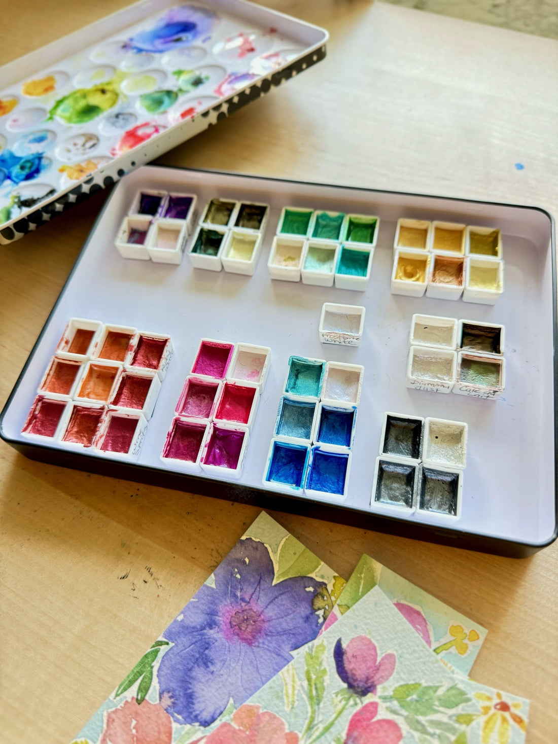 Watercolor palette with organized paint pans in various colors on a tray, with floral watercolor artwork partially visible below.