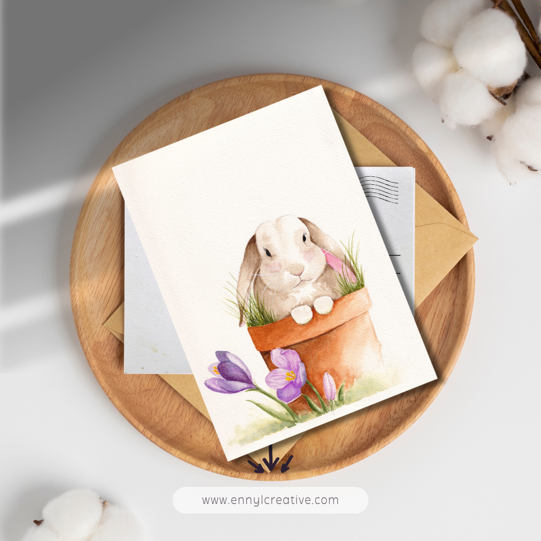Watercolor bunny in a flower pot with purple flowers, displayed as a greeting card on a wooden tray with envelopes.